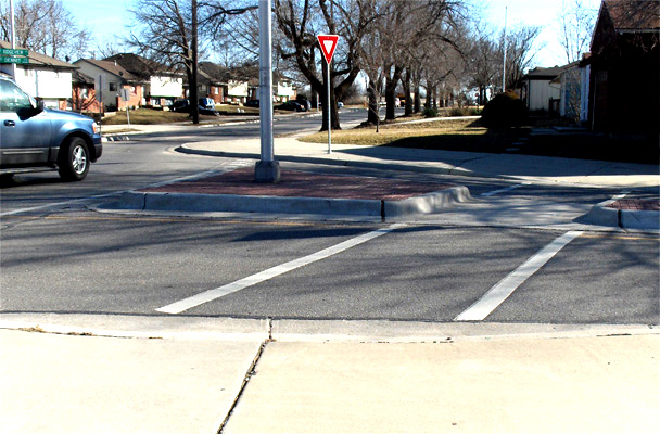 photo shows a street to the right of a circular lane.  The street has one narrow lane on each side of a refuge island that is about a half lane wide (the refuge island is triangular in shape, and widens to the left of the crosswalk and becomes more narrow to the right of the crosswalk